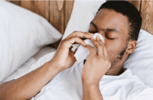 Black Man Blowing Runny Nose In Paper Tissue Having Rhinitis Lying In Bed At Home.